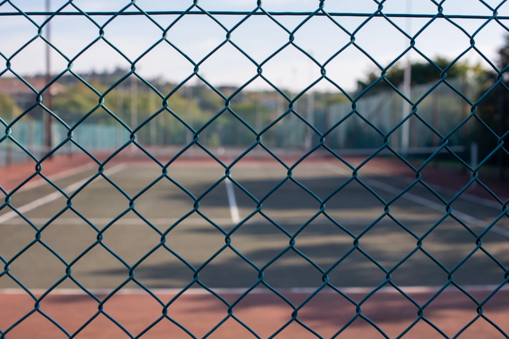 Ogrodzenie z siatki ogniowej w pierwszym planie, w tle pusty kort tenisowy z widocznymi liniami rozgraniczającymi