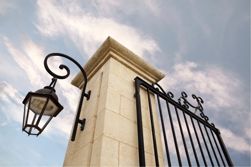 Słupek z jasnego kamienia i czarna, kuta brama wjazdowa z lampą zewnętrzną Bramy wjazdowe